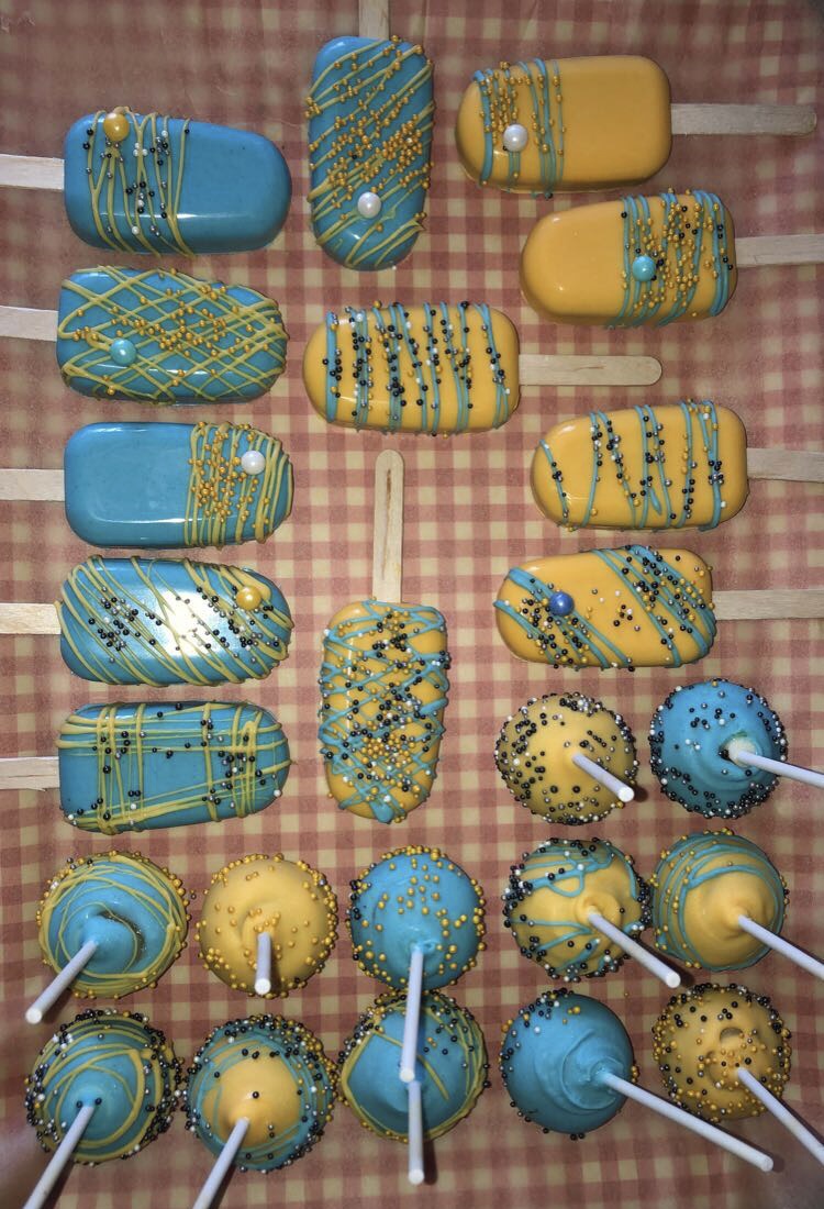 A red/white checkered tray of blue and yellow cakesicles and cake pops drizzled with blue and yellow icing and sprinkles.